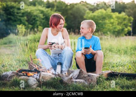 Lächelnde Schwester- und Bruderkinder unterhalten sich am Lagerfeuer fröhlich. Sie schauen sich gegenseitig Auge in Auge an, trinken Tee aus Thermoskannen. Frohe Familie Stockfoto