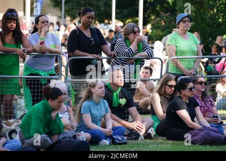 Auf einer Großleinwand am Fuß des Grenfell Tower in London beobachten die Menschen eine Zeremonie zur Festlegung von Glaubensbekenntnis und Kranz, um an die zu erinnern, die am 14 2018. Juni beim Feuer im Grenfell Tower ums Leben kamen. Bilddatum: Dienstag, 14. Juni 2022. Stockfoto