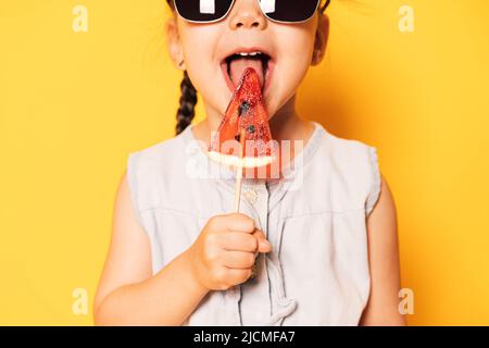 Mädchen in Sonnenbrillen leckt Lollipop in Form von Stück Wassermelone vor gelbem Hintergrund Stockfoto