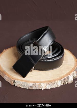 Aufgerollter schwarzer modischer Herren Ledergürtel mit automatischer Schnalle auf Holzscheibe auf braunem Hintergrund. Männliches Zubehör. Vorderansicht, vertikales Foto. Stockfoto