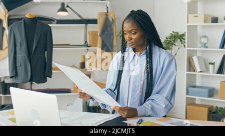 Ein talentierter afroamerikanischer Modedesigner kreiert eine neue Modekollektion, während er in einem Fashion Business Startup Studio an einem Laptop arbeitet. Konzept Stockfoto