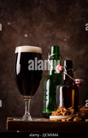 Glas dunkles Bier mit Schaumstoffkopf auf dunklem Holz Hintergrund mit leeren Flaschen und Biersnacks Stockfoto