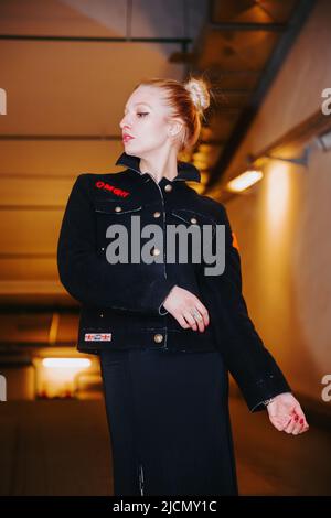 Das hübsche blonde Mädchen mit Haarsträhne, das einen langen schwarzen Mantel trägt, steht unter der Erde in der Nähe des Eingangs und Ausgangs einer Parkgarage; sie sieht seitlich aus Stockfoto