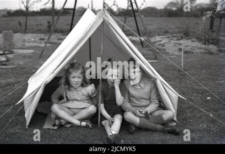 1950s, historisch, draußen auf einer Rasenfläche eines Spielplatzes, drei kleine Kinder sitzen nebeneinander in einem Zeltdach der damaligen Zeit, England, Großbritannien. Stockfoto