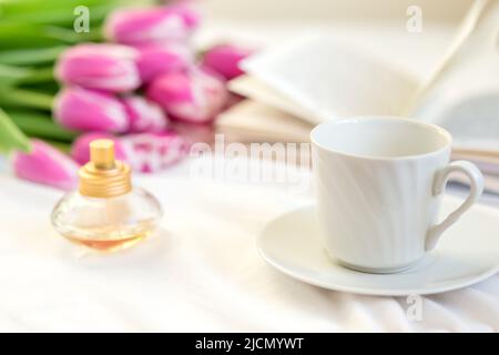 Ein Stillleben mit einem weißen Becher, einer Flasche Parfüm, einem offenen Buch und einem Bouquet von schönen rosa Tulpen Stockfoto