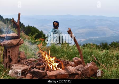 Nacht Sommer Camping am Seeufer. Eine Gruppe von fünf jungen, fröhlichen Touristen, die in hohem Gras am Lagerfeuer in der Nähe des Zeltes unter dem wunderschönen blauen Abendhimmel sitzen Stockfoto