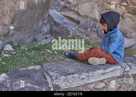 Kleiner Latino-Junge, der auf einer Wand sitzt und mit einer Kapuze über dem Kopf nach unten blickt, sehr nachdenklich. Stockfoto