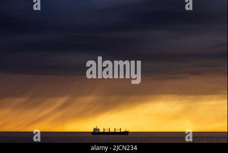 Rostock, Deutschland. 14.. Juni 2022. Ein Frachtschiff liegt auf den Straßen vor dem Hafen von Rostock. Am Horizont ziehen Regenwolken vor der tiefen Abendsonne über die Ostsee. Quelle: Jens Büttner/dpa/Alamy Live News Stockfoto