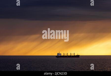 Rostock, Deutschland. 14.. Juni 2022. Ein Frachtschiff liegt auf den Straßen vor dem Hafen von Rostock. Am Horizont ziehen Regenwolken vor der tiefen Abendsonne über die Ostsee. Quelle: Jens Büttner/dpa/Alamy Live News Stockfoto