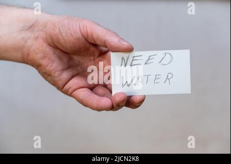 Ich brauche Wasser. Wörter in gezackten Buchstaben. Die Hand eines Mannes hält ein weißes Papierrechteck mit Text vor einem grauen Hintergrund. Stockfoto