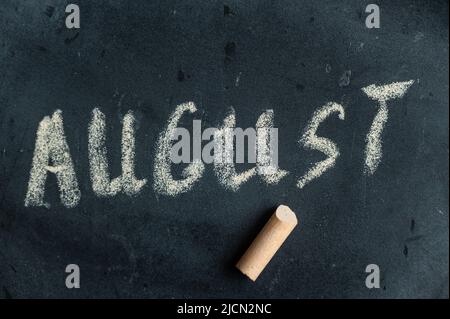 August. Name des letzten Sommermonats in gelber Kreide auf schwarzer Tafel geschrieben. Handgeschriebener Text. Daneben liegt ein Stück farbige Kreide. Stockfoto