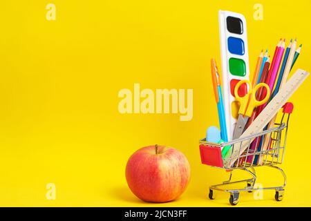 Apfel, Schreibwaren und Schulbedarf im Einkaufswagen, gelber Hintergrund Stockfoto