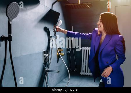 Elegante Verkäuferin in einem lila Anzug testet einen Duschkopf, Wasser läuft Stockfoto