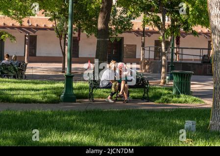 Im Santa Fe Plaza Park, Santa Fe, New Mexico, treffen sich zwei Frauen auf einer Parkbank, während sie über ein Telefon sprechen. Stockfoto