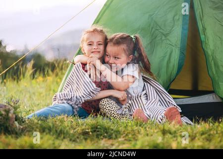 Zwei kleine umarmende Mädchen Schwestern sitzen auf dem grünen Gras neben dem Zelteingang und lächeln fröhlich. Sorglose Kindheit, Familienwerte und Outdoor Stockfoto