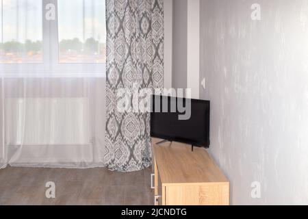 Heller Raum mit einem Vorhaufenfenster, einem Schrank mit einem Fernseher in der Nähe einer grauen Wand. Interieur eines kleinen Wohnzimmers, Schlafzimmer. Stockfoto