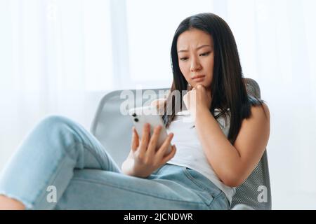 Verärgert verwirrt gebräunt schöne junge asiatische Frau lesen schockiert Nachricht mit Telefon zu Hause innen Wohnzimmer. Fernkommunikation, Fernarbeiten Stockfoto