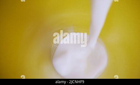 Die Milch wird in ein Glas aus der Nähe gegossen. Milch in einem Glas auf gelbem Hintergrund. Ein Blick von oben auf ein Glas Magermilch mit teilweise gefressenen Keksen Stockfoto