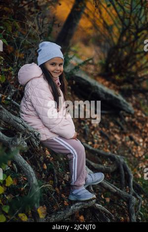 Junges kleines Mädchen, das auf Baumwurzeln im Wald sitzt, lächelt, die Kamera anschaut und mit Bäumen im Hintergrund posiert, Hut, rosa Jacke, Sport trägt Stockfoto