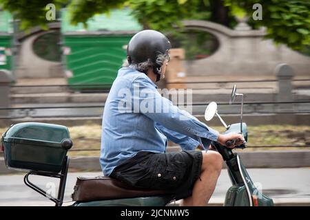 Belgrad, Serbien - 28. Mai 2022: Mann mittleren Alters auf einer grünen vespa-Scooter mit Heckkoffer auf der Stadtstraße neben dem Park Stockfoto