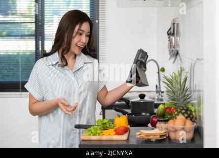 Fröhliche junge Vloggerin, die vom digitalen Tablet aus Videoanruf abruft, während sie zu Hause in der Küche kocht Stockfoto