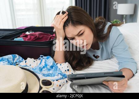 Gestresste Frau mit Tablet und Verpackung eines Koffers auf einem Bett zu Hause, Urlaub Reisekonzept Stockfoto