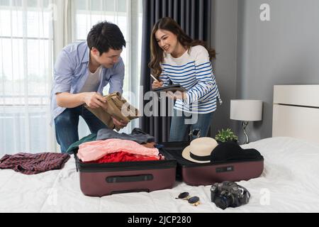 Ein Paar prüft eine Liste der Dinge mit einem Tablet, bereitet Kleidung vor und verpackt sie in Koffer auf einem Bett, um einen Reiseurlaub zu planen. Stockfoto