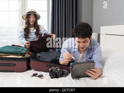 Ein Paar prüft eine Liste der Dinge mit einem Tablet, bereitet Kleidung vor und verpackt sie in Koffer auf einem Bett, um einen Reiseurlaub zu planen. Stockfoto