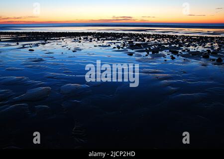 Die Sonne geht über Meer und Meer auf und spiegelt sich in den gewellten Sandmustern am Strand und an der Küste wider Stockfoto