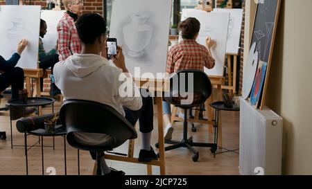 Künstlerische Person, die Foto von Vase Meisterwerk auf Leinwand. Schüler der Kunstklasse mit dem Smartphone, um nach dem Zeichnen des Modells eine Skizze zu fotografieren und so ein zeitgenössisches Meisterwerk zu retten. Stockfoto