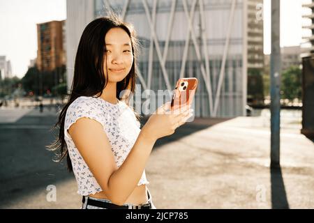 Junge asiatische Frau, die auf die Kamera schaut, während sie ihr Mobiltelefon auf der Straße im Freien benutzt. Stockfoto