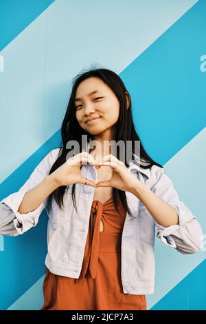 Porträt einer jungen asiatischen Frau, die die Kamera anschaut, während sie mit ihren Händen eine Herzform bildet. Stockfoto