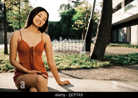 Junge asiatische Frau lächelt die Kamera an, während sie sich auf einer Bank im Freien entspannt. Stockfoto