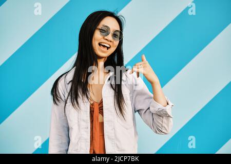Fröhliche junge asiatische Frau in Sonnenbrille lächelt, während sie vor einer gestreiften Wand posiert. Stockfoto