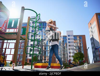 Glückliche blonde Mutter fangen fröhliche Tochter, Springen von der Leiter auf dem Spielplatz. Schöne Frau, die Spaß mit einem weiblichen Kind im modernen Innenhof der Stadt Wohnhochhäuser hat. Stockfoto