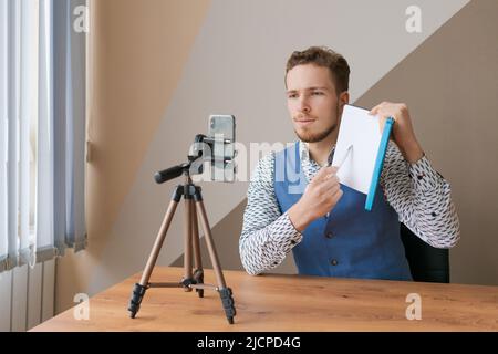 Lächelnder Geschäftsmann mit Telefon und Stativ, macht Videoanruf für Kollegen, sitzt im Büro am Tisch, soziales Online-Netzwerk, Videoblogger im mittleren Alter. Stockfoto