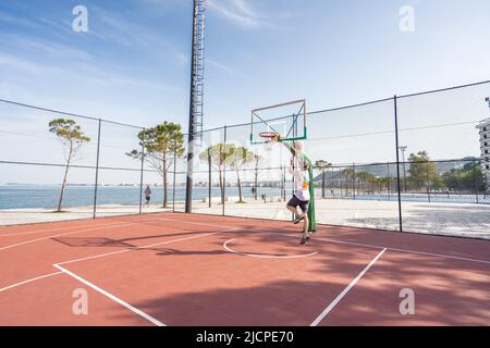 Vlore, ALBANIEN - 20. MAI 2022: Männlicher Basketballspieler schießt einen Sprung im Freien. Allein Ball spielen. Sonnenaufgang Ozean Hintergrund. Stockfoto