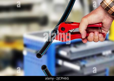 Handwerkswerkzeuge. Ein Mann schneidet ein Stück PE-Druckrohr oder Wasserrohr mit einem roten pvc-Rohrschneider über abstrakten industriellen Hintergrund genau. Trinken Stockfoto