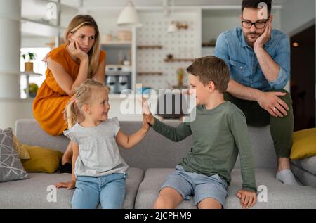 Müde Eltern, die auf der Couch sitzen, fühlen sich erschöpft genervt, während glückliche Kinder zusammen spielen. Stockfoto