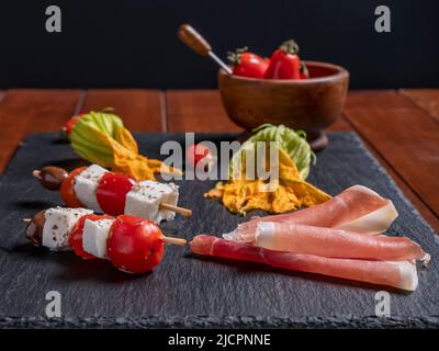 Zwei Spieße mit Käsestücken und Kirschtomaten auf einem Schieferteller, daneben Zucchini-Blumen und Schinken in Scheiben Stockfoto