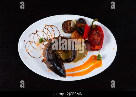 Weißer Teller mit gegrillter Forelle mit Kartoffeln, rotem Pfeffer, Tomaten und Zitrone auf schwarzem Hintergrund Stockfoto