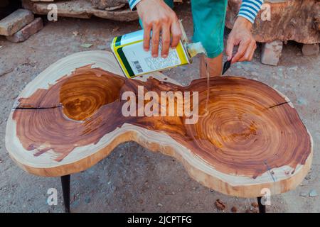 Zimmermann gießt Leinöl auf einen Holztisch. Prozess der Herstellung von Holztisch Stockfoto