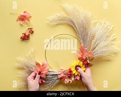Hände, die getrockneten Blumenkranz aus trockenem Pampagras und Herbstblättern machen. Flach auf gelbem Papier, Sonnenlicht mit langen Schatten. Stockfoto