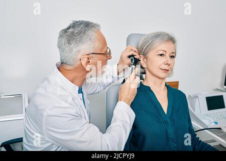 Der Arzt des Otolaryngologen überprüft das Ohr der älteren Frau mit einem Otoskop oder einem Ohrhörer in der Hörzentrale. Audiologie Stockfoto