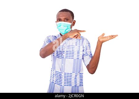 Ein junger Mann in einem Hemd, das auf weißem Hintergrund steht und mit der medizinischen Maske mit Hand und Finger zeigt. Stockfoto