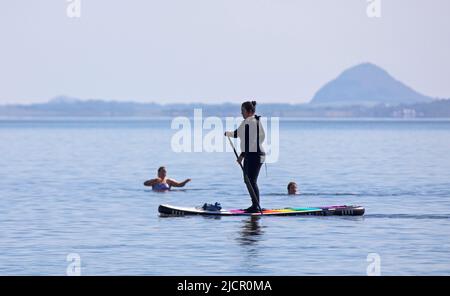 Portobello, Edinburgh, Schottland, Großbritannien. 15.06.2022. Hitzewelle verlockend die Paddlebarder auf den Firth of Forth, es ist ein paar Wochen her, dass sich irgendwelche aufgrund der jüngsten kühlen bewölkten Bedingungen gewagt haben. Die Temperatur von 18 Grad an diesem Morgen brachte einige für die Übung heraus. Im Bild: Paddlebarderin und Kaltwasserschwimmerin mit Berwick Law im Hintergrund. Kredit: Scottiscreative/alamy Live Nachrichten. Stockfoto