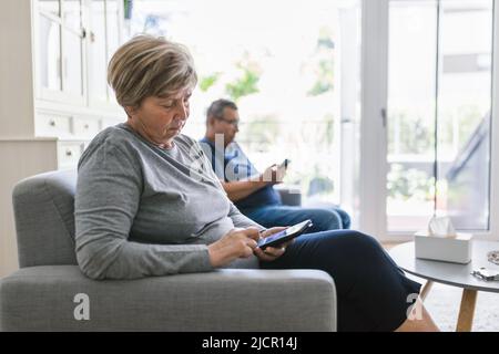 Ein älteres Paar sitzt auf der Couch und benutzt zu Hause ihr Mobiltelefon Stockfoto