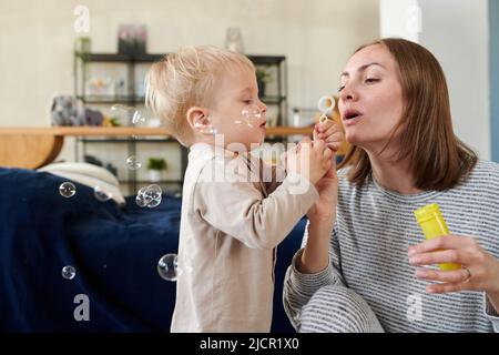 Junge Mutter bläst Seifenblasen, während ihr kleiner Sohn versucht, sie während ihrer Spiele zu Hause zu fangen Stockfoto