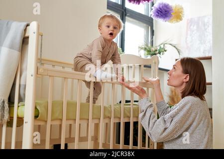 Das kleine Kind wacht am Morgen auf und klettert aus seiner Krippe heraus, während die Mutter ihre Hände zu ihm streckte Stockfoto