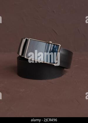 Aufgerollter schwarzer modischer Herren Ledergürtel mit automatischer Schnalle auf braunem Hintergrund. Männliches Zubehör. Vorderansicht, vertikales Foto. Stockfoto